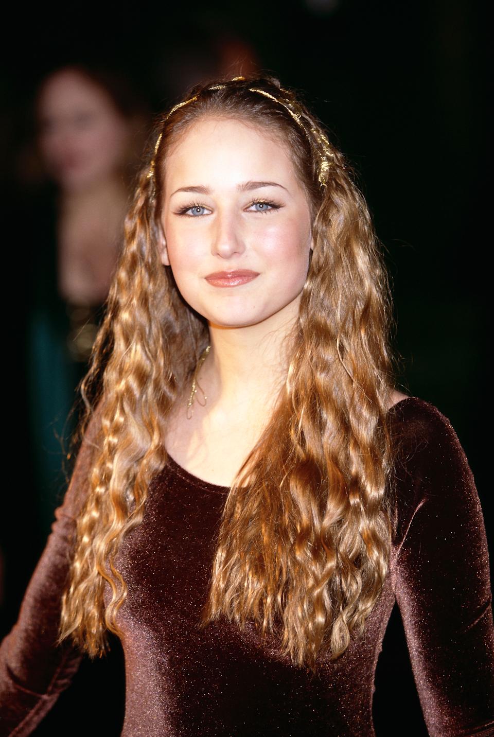 Uma pessoa com cabelos ondulados compridos é colocado em um vestido de veludo em um evento, sorrindo um pouco